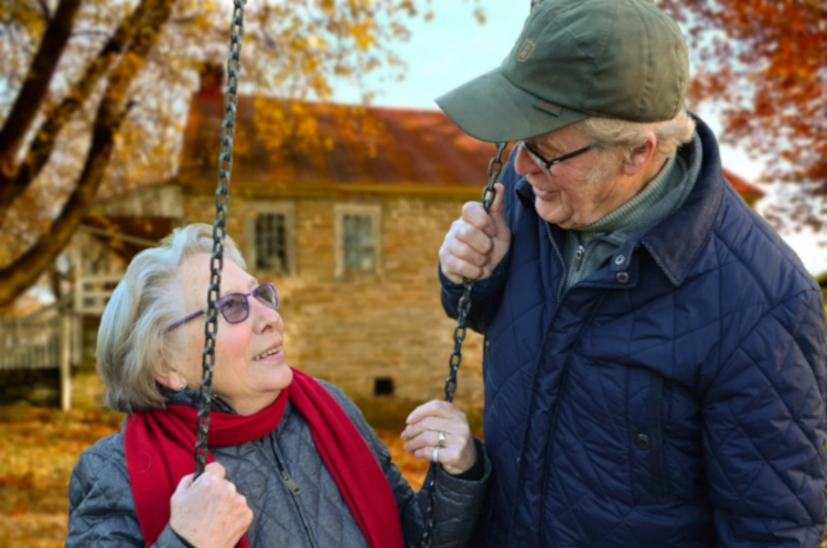 Couple de personnes âgées devant leur maison en pierres, Madame étant assise sur une balançoire, qui a utilisé la sophrologie , près de Rouen, pour bien vieillir et accompagner les changements qui se sont mis en place avec le vieillissement.