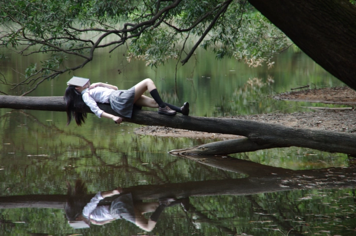 Une femme allongée sur une branche au dessus d'une rivière, se repose avec un livre posé sur la tête et se laisse aller comme elle a appris pendant les séances de sophrologie près de Rouen, elle qui n'arrivait pas à lâcher prise et qui était toujours dans le contrôle auparavant.